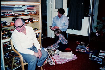 Vintage indoor scene featuring three generations: an older man in glasses and a white shirt seated, a woman in a plaid shirt ...