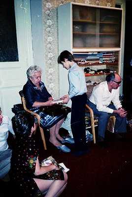 Vintage indoor scene featuring four individuals in a modestly furnished room, likely mid-20th century. A seated woman in trad...