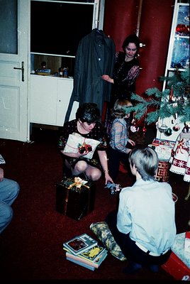 Vintage holiday scene featuring a family opening gifts under a decorated Christmas tree in a cozy 1970s living room. A woman ...