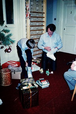 Family gathering in a Soviet-era apartment during holiday celebrations, likely late 1970s–early 1980s. A man and boy open a L...