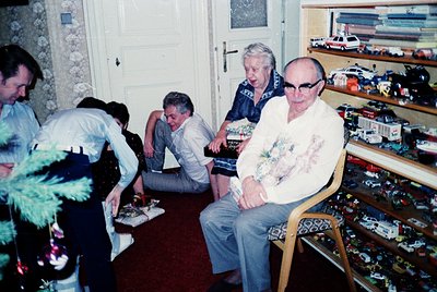 Vintage indoor scene featuring five men in a cluttered room with mid-century decor. Two seated men in casual attire (one in a...