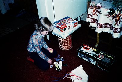 Child assembling a toy camera (Kenwood Carrera Turbo) on a patterned rug, surrounded by wrapped gifts and holiday-themed tabl...