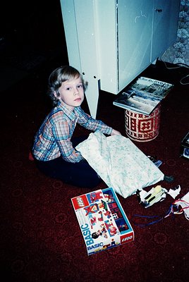 Child in plaid shirt sits on patterned carpet, examining a folded floral bedsheet beside an open Lego BASIC set and a woven b...