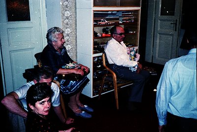 Vintage indoor gathering featuring four adults in a dimly lit room with wooden paneling and floral wallpaper. A man in sungla...