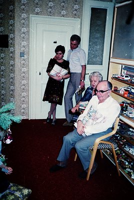 Vintage indoor portrait featuring three adults in a patterned, floral-wallpapered room. A seated man in a white shirt and pat...