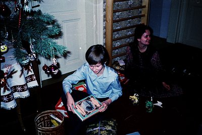 Vintage indoor scene featuring a young boy (1970s-80s) reading a book beside a decorated Christmas tree. Decor includes woven...