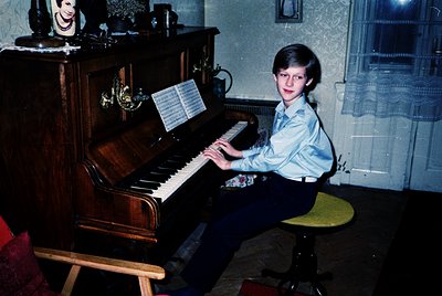Vintage indoor portrait of a boy playing a grand piano in a residential setting, likely 1970s–1980s. Wooden piano with ornate...