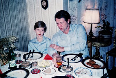 Vintage indoor family portrait featuring a man and boy seated at a set table with 1970s-era tableware. The man wears a light ...