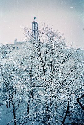 Vintage sepia-toned winter scene featuring snow-laden bare trees framing a tall, cylindrical tower with a spire, likely a chu...