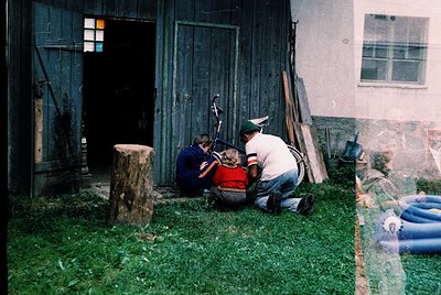 Vintage photo of two men tending to a small dog in a rural courtyard. Wooden barn with double doors and weathered planks; vis...