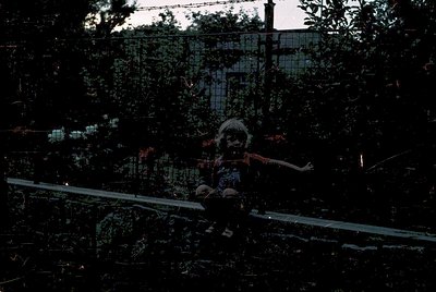Vintage black-and-white photo of a child sitting on a rusted metal bench in an abandoned urban area. Overgrown foliage and ba...