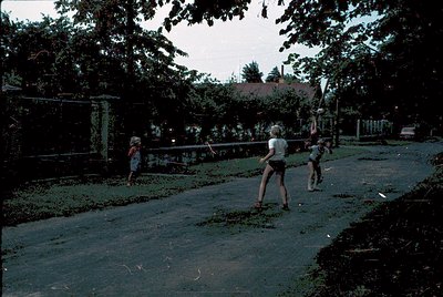 Vintage black-and-white photo of three children playing frisbee in a residential backyard, likely 1970s. Concrete pathway bor...