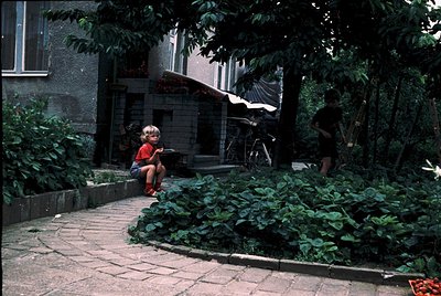 Vintage courtyard scene featuring two children in 1970s-era clothing—one in a red sweater and shorts, seated on a low wall; t...