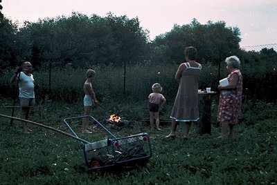 Family gathers around an outdoor fire in a rural setting, likely 1960s–1970s. Adults and children in casual summer attire—men...