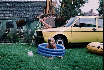 Vintage backyard scene featuring a yellow 1970s-era sedan (likely a Škoda) parked on grass. Two children play in an inflatabl...