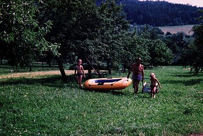 Family of four in a rural outdoor setting, mid-20th century. Adult leads two young children toward an orange inflatable raft ...
