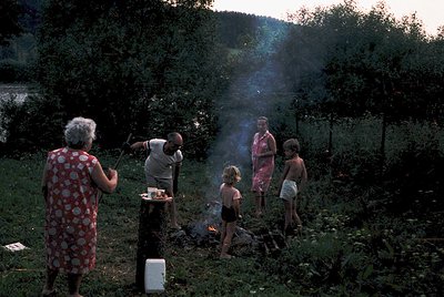 Family gathers around an outdoor fire pit in a rural setting, likely mid-20th century. Adults in casual summer attire (1960s-...