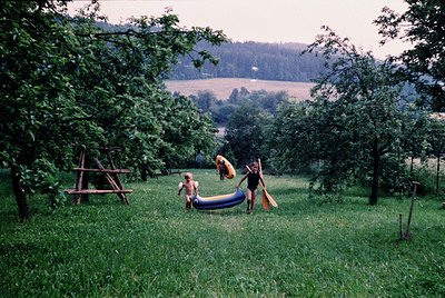 Vintage 1970s rural scene: two adults and a child playing with a blue inflatable slide in a lush orchard. Rolling hills and a...