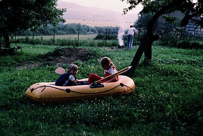 Two children in 1970s-style inflatable rafts on grassy rural terrain, likely Eastern Europe. Adult in background tending to a...