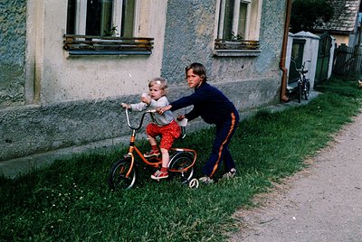 A woman in 1970s-style track pants assists a toddler on a small orange bicycle beside a concrete residential building with wh...