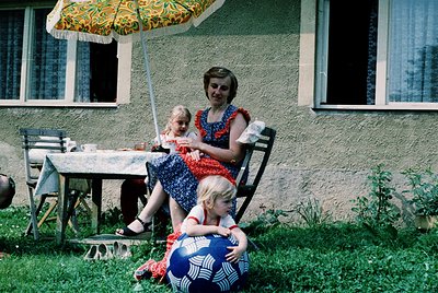 A woman in a 1960s floral-patterned dress and red collar sits on a wicker chair under a yellow floral umbrella, cradling a yo...