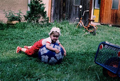 Vintage childhood scene: young child in 1970s-style patterned dress and hat plays with a blue exercise ball on overgrown gras...