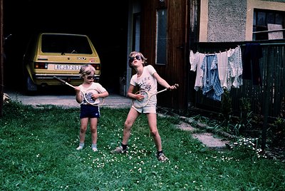 Two children in 1970s-style swimwear play with tennis rackets in a residential backyard. The older child wears a white tank t...