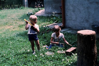 Two children play outdoors with homemade toys in a grassy backyard, likely mid-20th century. The boy on left holds a yellow-h...