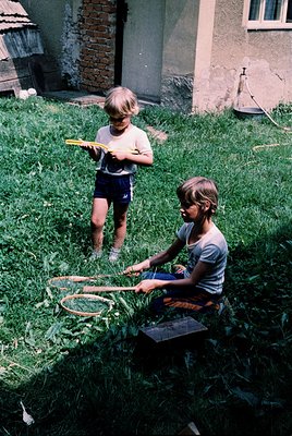 Two children play with homemade badminton rackets in a grassy backyard, 1960s-1970s. Brick house with exposed chimney and woo...