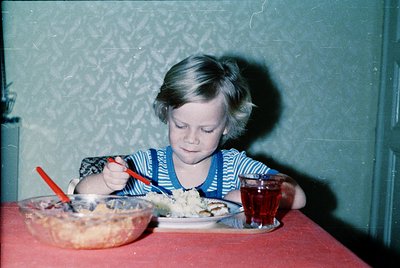Vintage indoor portrait of a young child eating cake at a table, wearing a striped shirt and bib. Red plastic fork and glass ...