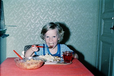 Vintage indoor portrait of a child mid-meal, seated at a red table with a striped apron. Plate holds cake with frosting, plas...