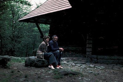 Two individuals sit on a rock beneath a rustic wooden shelter in a forested area, likely mid-20th century. The man wears a st...