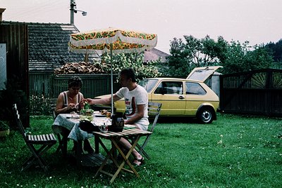 Two individuals enjoy a meal outdoors under a patterned umbrella, seated at a foldable table. The man wears a vintage T-shirt...