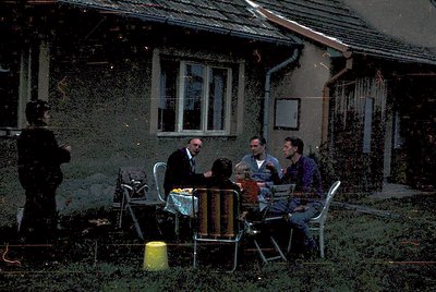 Vintage outdoor gathering at a weathered concrete building, likely 1970s Eastern Europe. Four adults seated at a metal table ...