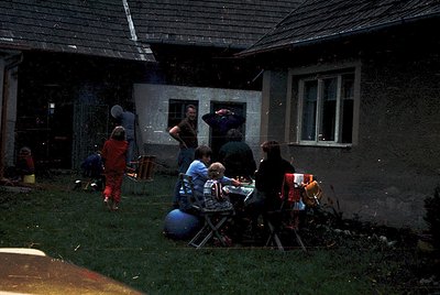 Vintage backyard gathering in a modest, single-story home with concrete walls and a sloped roof. Six adults and children seat...