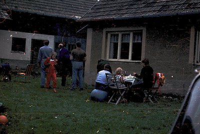 A mid-20th-century backyard gathering in a Soviet-era residential block. Six adults and children in casual 1970s attire—plaid...
