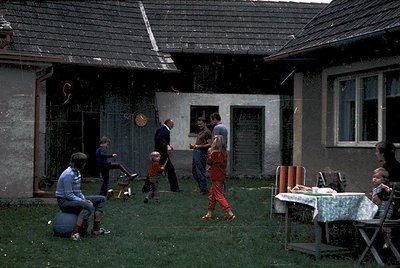 Vintage mid-century courtyard scene with concrete block house and grassy yard. Adults and children play casually—one child in...