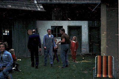 Vintage group photo outside a weathered, Soviet-era concrete building with graffiti. Five adults in 1970s workwear (overalls,...