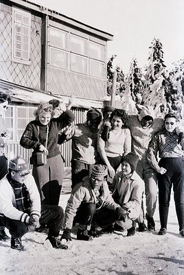 Group of 10 people posing in winter attire outside a wooden building with snow-covered ground, likely a ski resort. Mid-20th ...
