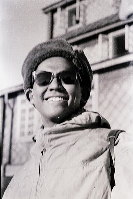 Black-and-white portrait of a person wearing a beret, round sunglasses, and a draped jacket over a shoulder. Urban background...