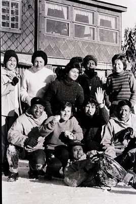 Vintage black-and-white group photo of 8 people posing outdoors in winter attire, likely –. Snow-covered ground and a brick b...