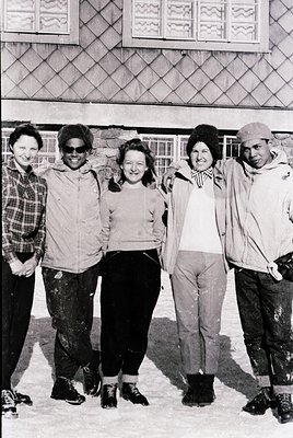 Five individuals pose in winter attire on snow-covered ground, likely a ski resort or urban plaza. Distinctive elements: plai...