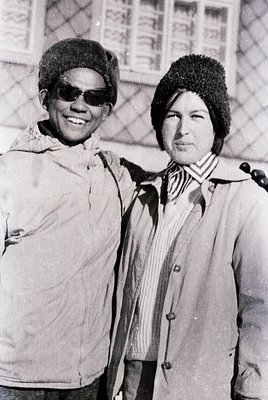Two individuals pose outdoors in 1960s-style attire—men’s sunglasses, knit caps, and buttoned coats—against a patterned brick...