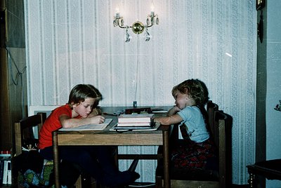 Two children studying at a wooden table in a dimly lit room, likely mid-1980s. The boy in red focuses on a notebook, while th...