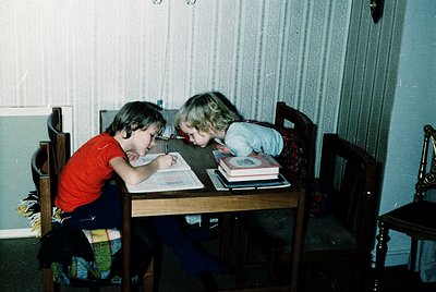 Two children engrossed in reading at a wooden table in a vintage interior, likely mid-1980s–1990s. The boy wears a red shirt ...