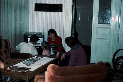Vintage indoor scene featuring a family group around a table with a retro TV (likely 1970s-80s). Adults and children gather i...