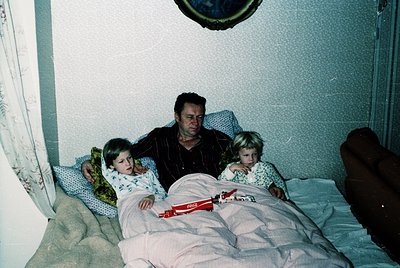 Family portrait in a modest bedroom, likely mid-to-late 20th century. A man sits between two children on a double bed with a ...