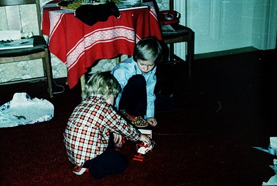 Two children play with a toy train set in a vintage indoor setting. The girl in a red-and-white plaid shirt kneels, while the...