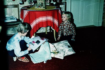 Two children in 1970s-style clothing examine wrapped gifts under a festive tablecloth with holiday ornaments. The boy kneels,...