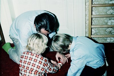 A tender moment captured indoors, featuring an adult kneeling beside two young children on a patterned rug. The adult, dresse...